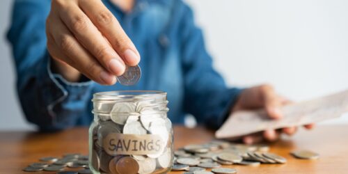 1185358443_The woman hand is putting a coin in a glass bottle and a pile of coins on a brown wooden table,Investment business, retirement, finance and saving money for future concept. -min